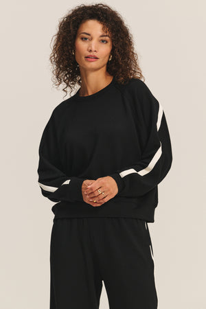 A woman with curly hair wears the FRANKIE SWEATSHIRT by Velvet by Graham & Spencer, featuring white sleeve stripes, paired with matching black pants against a plain background.