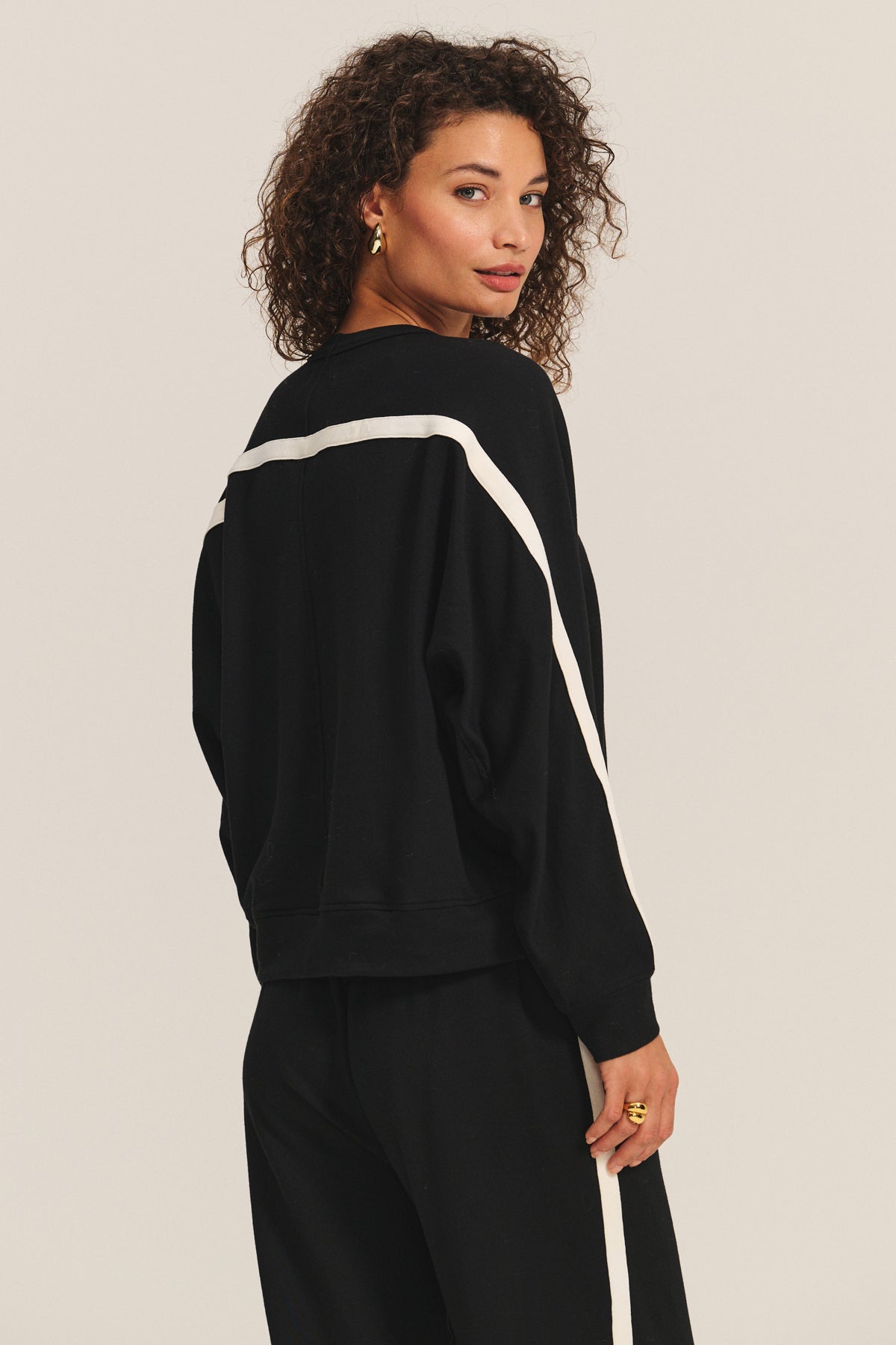 A woman with curly hair models the FRANKIE SWEATSHIRT by Velvet by Graham & Spencer, featuring black fabric with white stripes and relaxed dolman sleeves, paired with matching black pants, as she looks over her shoulder against a plain background.-Hover Image