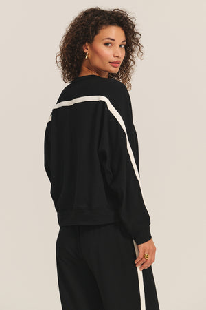 A woman with curly hair models the FRANKIE SWEATSHIRT by Velvet by Graham & Spencer, featuring black fabric with white stripes and relaxed dolman sleeves, paired with matching black pants, as she looks over her shoulder against a plain background.