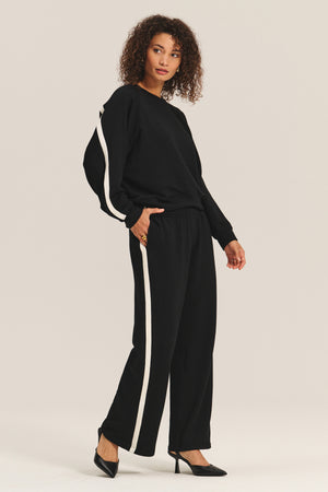 A woman wears the FRANKIE SWEATSHIRT by Velvet by Graham & Spencer, styled with wide-leg, white-striped pants and black heels, standing against a plain background.