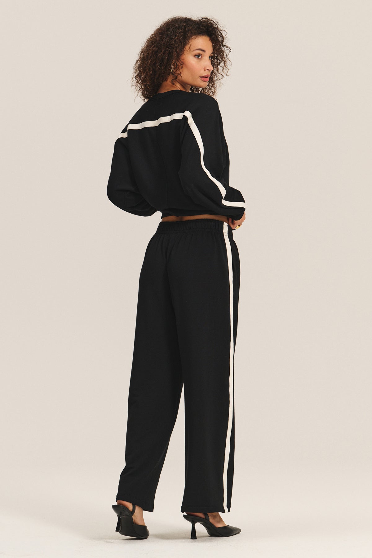 A person wears the FRANKIE SWEATSHIRT by Velvet by Graham & Spencer with relaxed dolman sleeves, paired with matching black pants featuring white stripes and black heels, standing and looking over their shoulder.-40247639081153