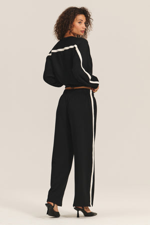 A person wears the FRANKIE SWEATSHIRT by Velvet by Graham & Spencer with relaxed dolman sleeves, paired with matching black pants featuring white stripes and black heels, standing and looking over their shoulder.