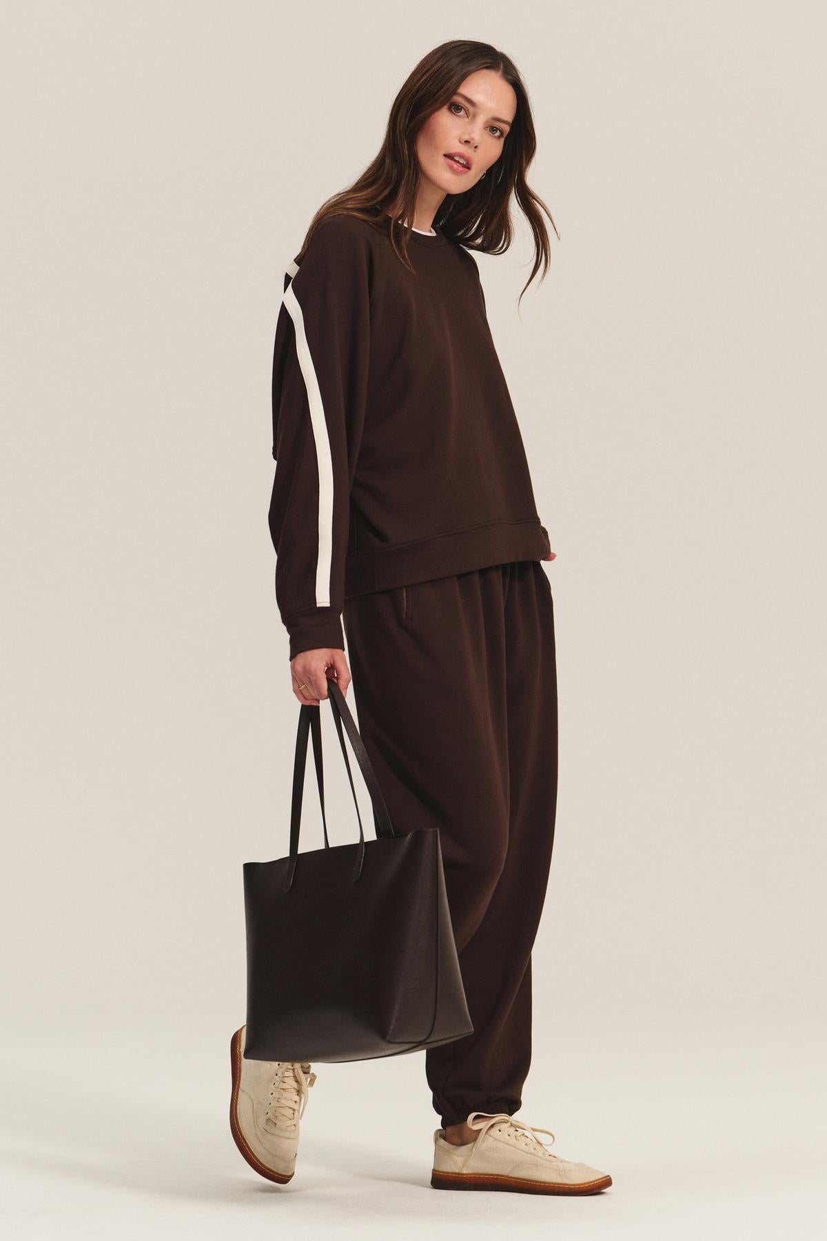 Woman wearing the Velvet by Graham & Spencer EVERS JOGGER, a relaxed fit brown tracksuit with white stripes and elastic waist, paired with beige sneakers and a large black tote bag, standing against a neutral background.-40626638094529