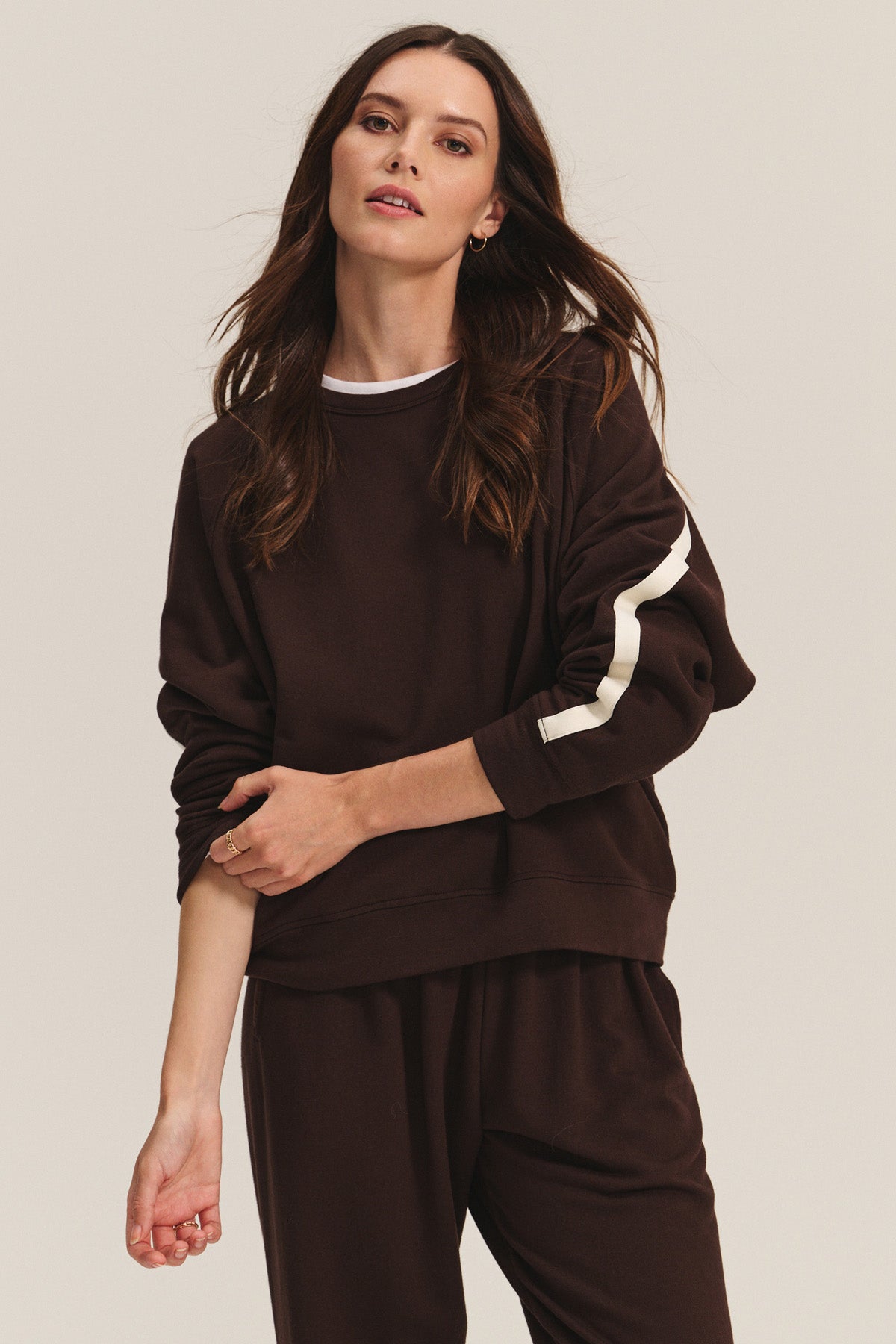 A woman wears the FRANKIE SWEATSHIRT by Velvet by Graham & Spencer, featuring relaxed dolman sleeves and a white sleeve stripe, paired with matching pants against a plain background.