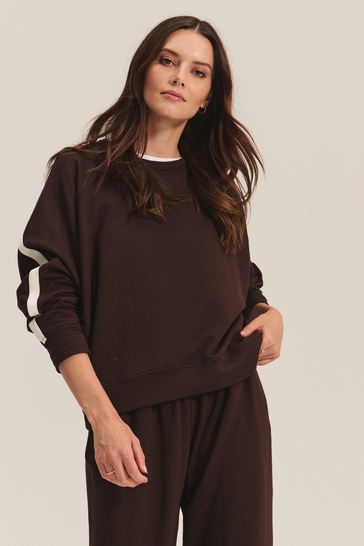 A woman wears the FRANKIE SWEATSHIRT by Velvet by Graham & Spencer—a brown midweight fleece with white sleeve stripes—paired with matching brown pants, standing against a plain background.