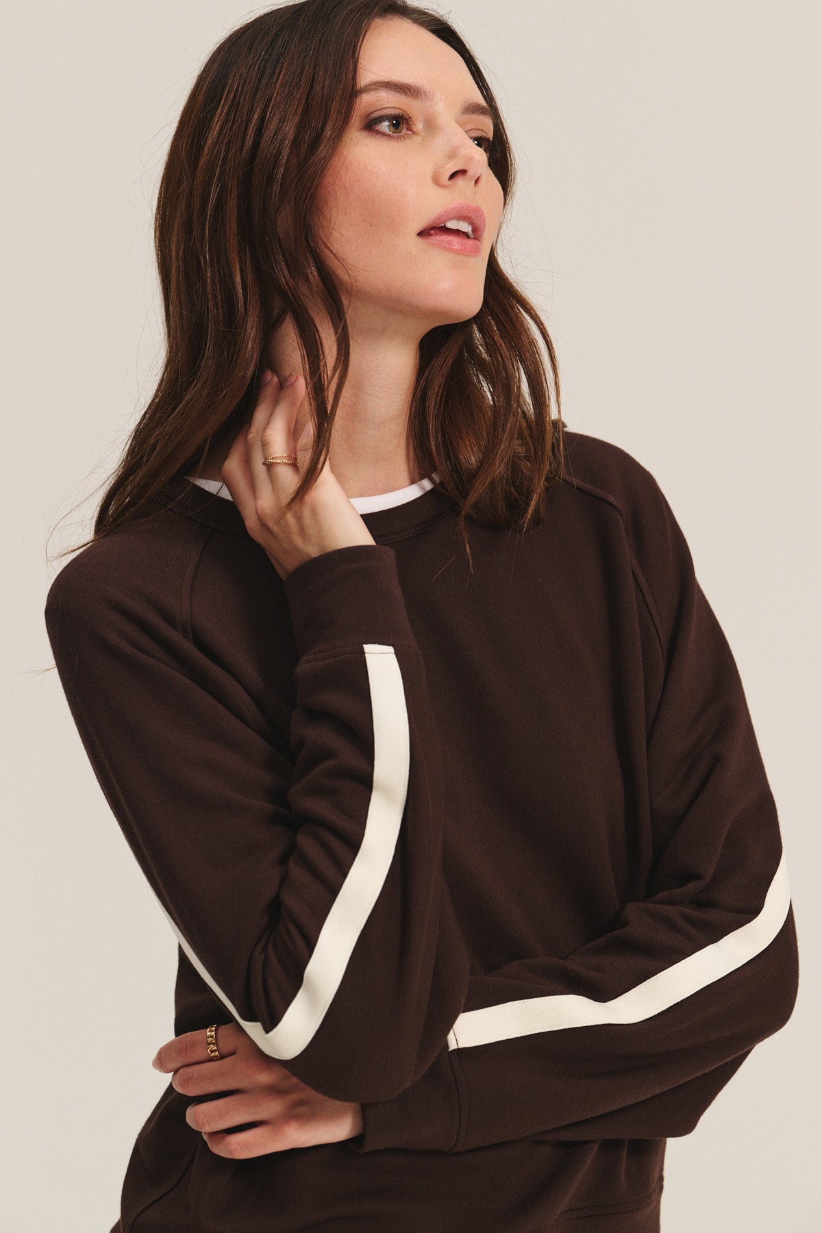 A woman models the FRANKIE SWEATSHIRT by Velvet by Graham & Spencer—a brown long-sleeve top with white sleeve stripes—glancing sideways, her hand to her neck, highlighting its modern, sporty comfort.