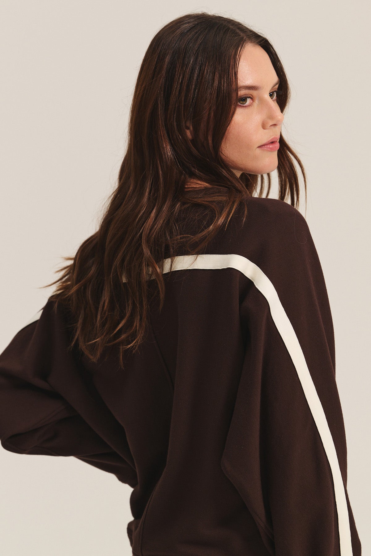 A woman with long brown hair wears the FRANKIE SWEATSHIRT by Velvet by Graham & Spencer in dark brown with a white shoulder stripe, looking over her left shoulder against a plain background.-40598482059457