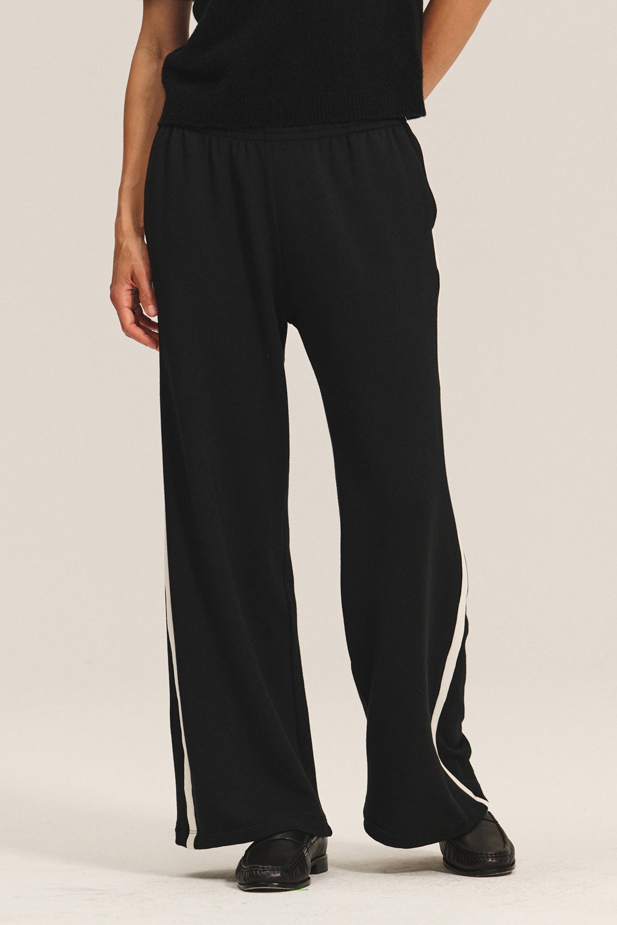   Wearing the Velvet by Graham & Spencer GIGI SWEATPANT, a person in relaxed fit black pants with white side stripes, paired with a black top and shoes, stands against a plain background. 