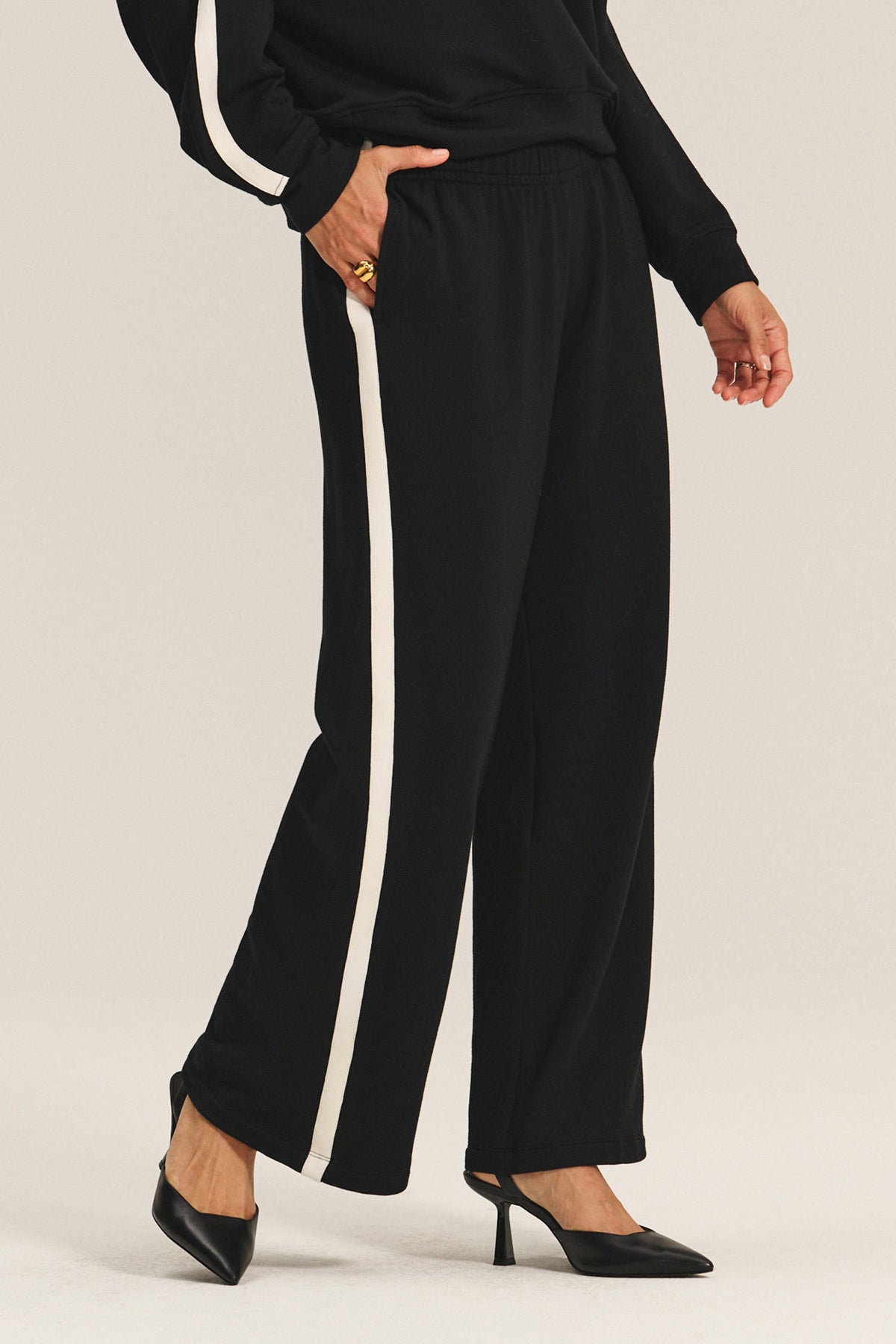   A person wearing the GIGI SWEATPANT by Velvet by Graham & Spencer—black relaxed fit pants with a white side stripe—and black high-heeled shoes, shown from shoulder to feet. 