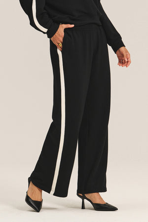 A person wearing the GIGI SWEATPANT by Velvet by Graham & Spencer—black relaxed fit pants with a white side stripe—and black high-heeled shoes, shown from shoulder to feet.