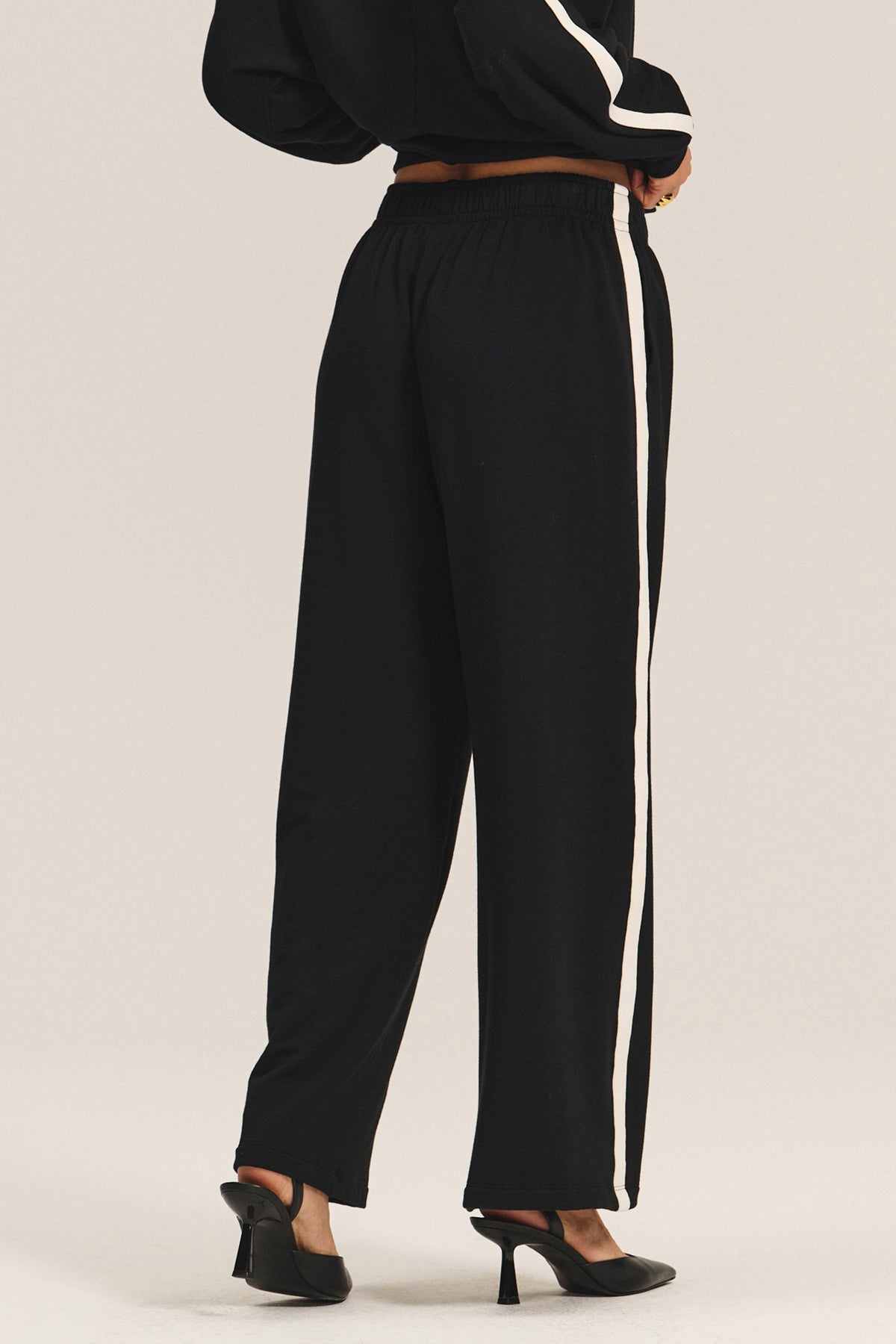   Model wears the GIGI SWEATPANT by Velvet by Graham & Spencer—a relaxed fit, black wide-leg sweatpant with a white side stripe—styled with black heels and photographed from the back against a plain light background. 