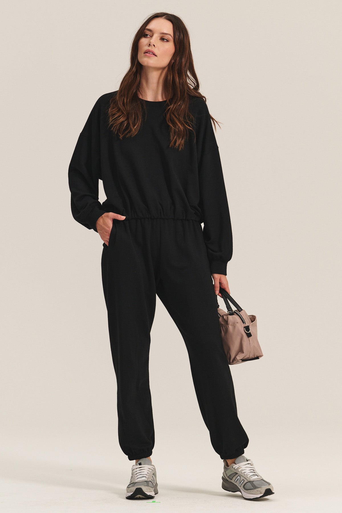 A woman wears the Velvet by Graham & Spencer EVERS JOGGER in black with a relaxed fit, paired with gray sneakers and a small beige handbag, standing against a plain light background.