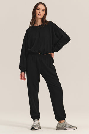A woman wears the EVERS JOGGER by Velvet by Graham & Spencer—relaxed fit black fleece joggers and a matching sweatshirt—paired with gray New Balance sneakers, standing against a plain light background.