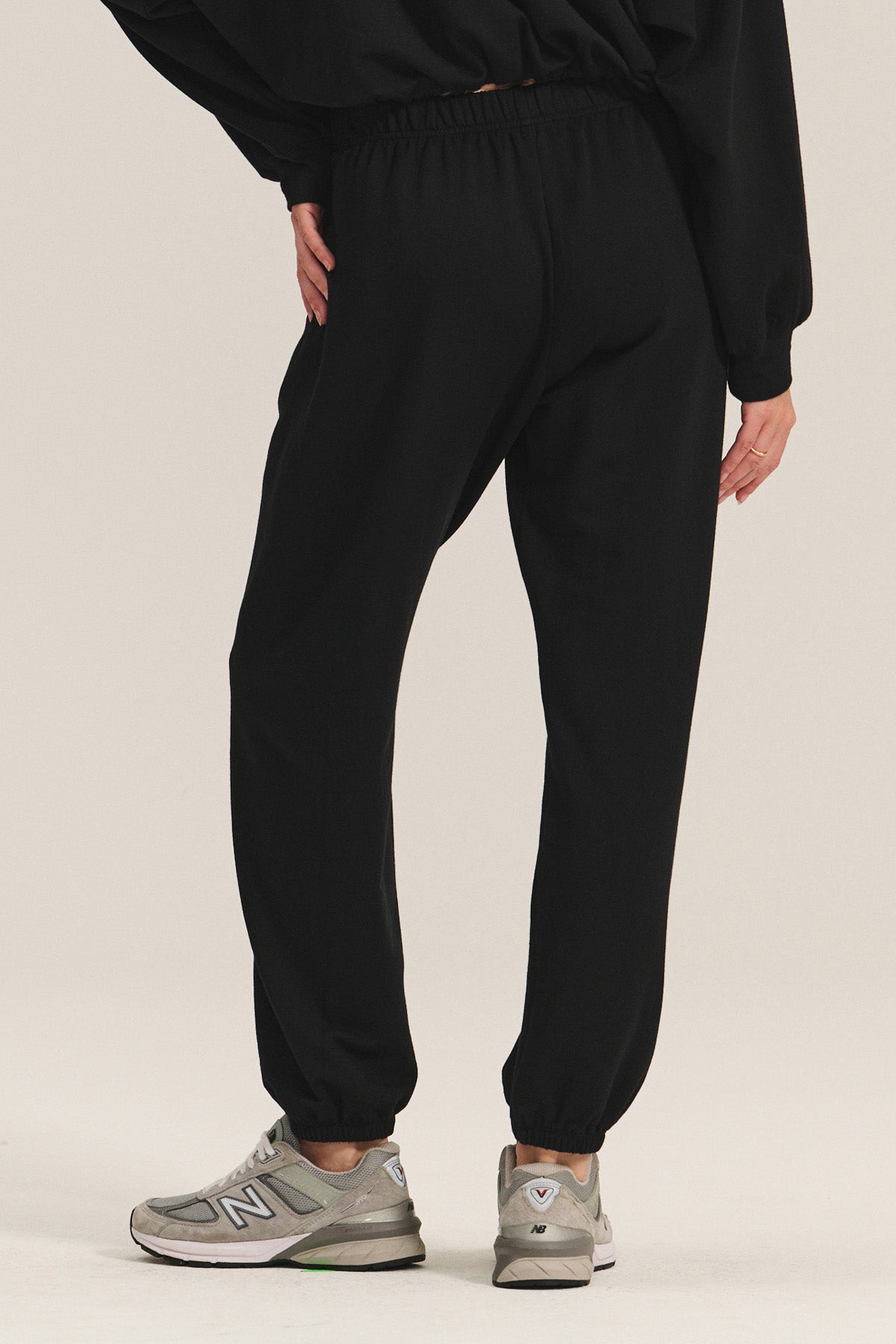 A person stands on a light background, shown from the back, wearing Velvet by Graham & Spencer’s EVERS JOGGER relaxed fit black fleece pants with an elastic waist, a black top, and gray New Balance sneakers.