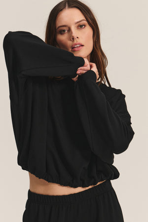 A woman models the JOLIE SWEATSHIRT by Velvet by Graham & Spencer—a loose-fitting black fleece top with an elastic bubble hem—and matching black pants, posing with one arm raised against a plain background.
