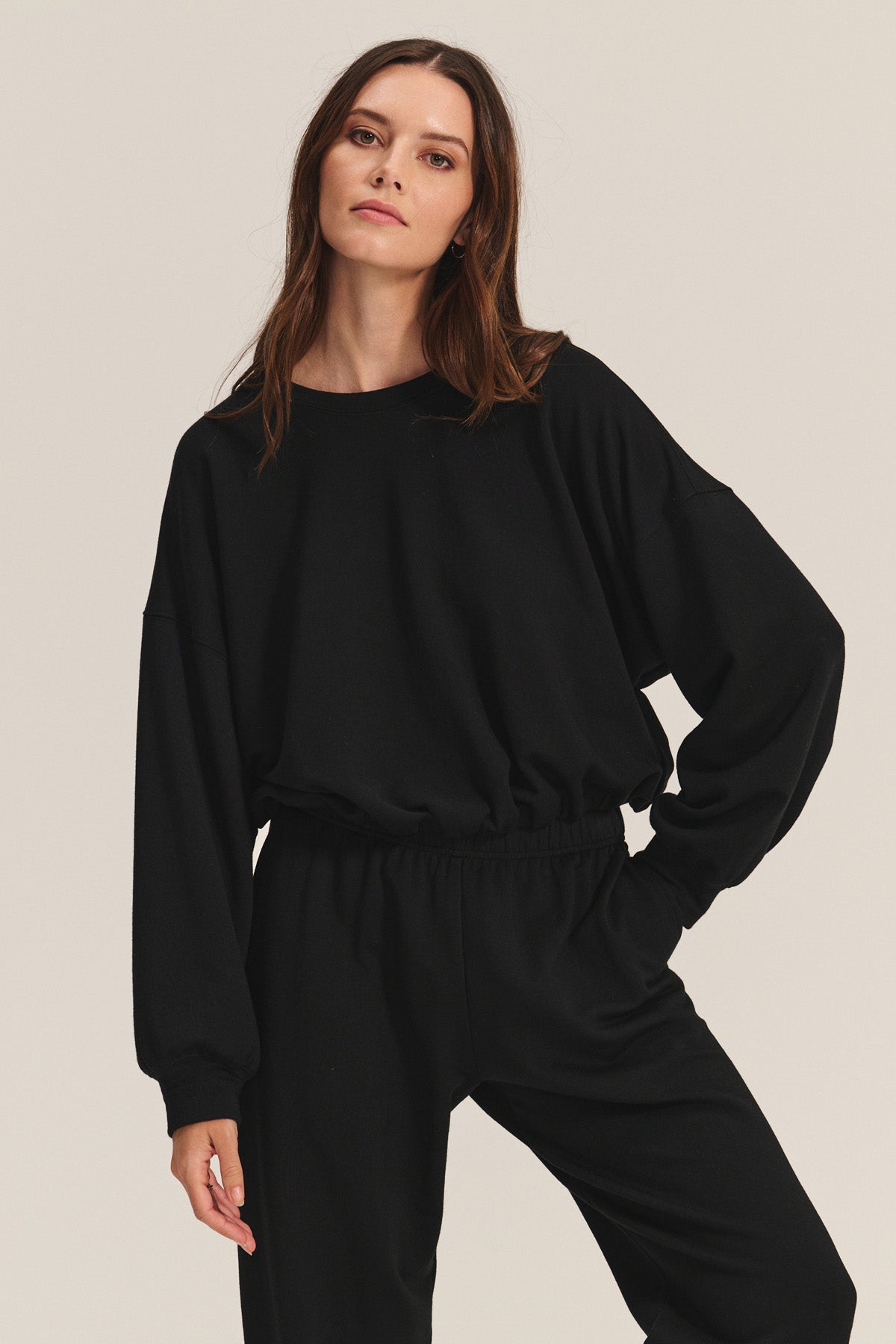A woman wears the JOLIE SWEATSHIRT by Velvet by Graham & Spencer— a loose black fleece top with an elastic bubble hem—paired with matching pants, posing with one hand on her hip against a plain background.
