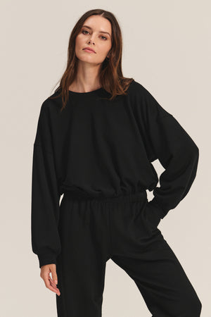 A woman wears the JOLIE SWEATSHIRT by Velvet by Graham & Spencer— a loose black fleece top with an elastic bubble hem—paired with matching pants, posing with one hand on her hip against a plain background.