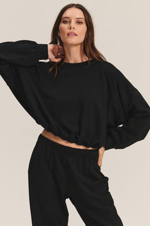 A person models the JOLIE SWEATSHIRT by Velvet by Graham & Spencer, a loose-fitting black fleece cropped top with an elastic bubble hem, paired with matching black pants against a plain background.