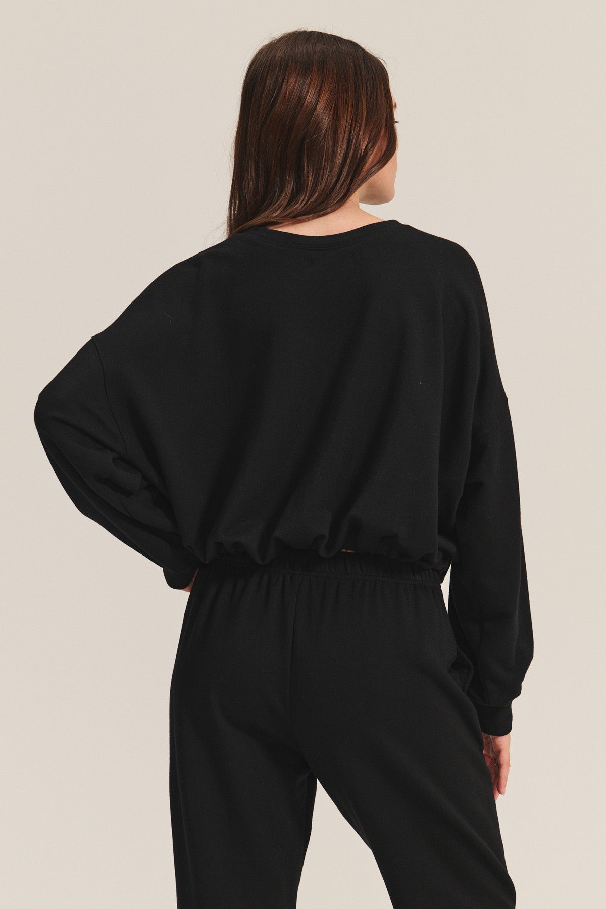 A person with long brown hair stands facing away against a plain light background, wearing the JOLIE SWEATSHIRT by Velvet by Graham & Spencer— a black fleece cropped sweatshirt with an elastic bubble hem—paired with black pants.-40334221803713