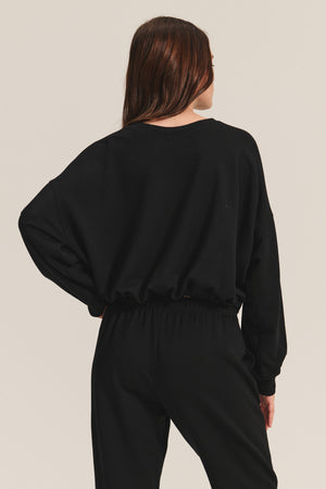 A person with long brown hair stands facing away against a plain light background, wearing the JOLIE SWEATSHIRT by Velvet by Graham & Spencer— a black fleece cropped sweatshirt with an elastic bubble hem—paired with black pants.