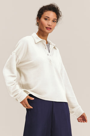 A person wears the MARIAN SWEATSHIRT by Velvet by Graham & Spencer—a cream, long-sleeve collared shirt with a relaxed fit—layered over a gray top and navy pants, standing against a plain background.