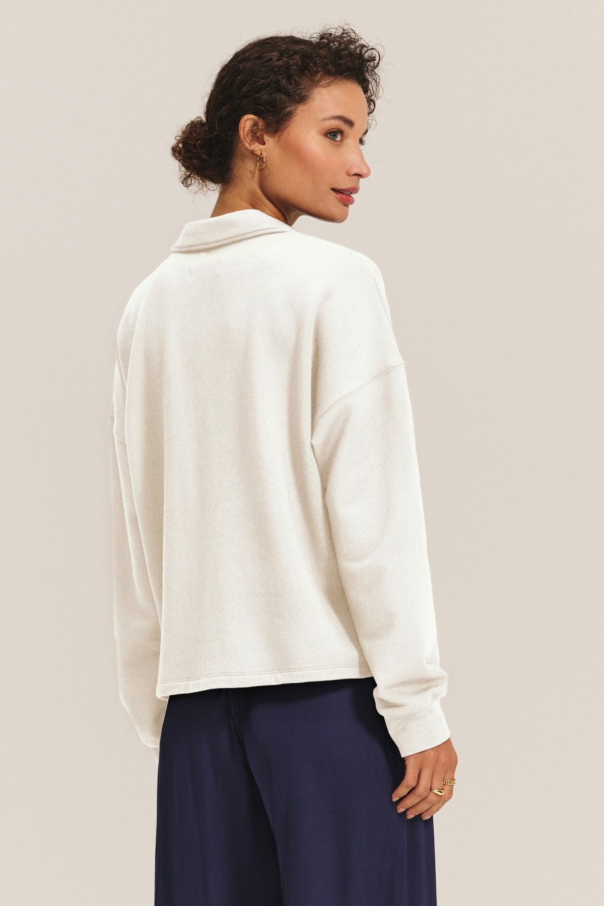 A woman faces away, showcasing the back of the relaxed MARIAN SWEATSHIRT by Velvet by Graham & Spencer in off-white, styled with dark navy pants against a plain light background.