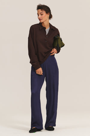 A person stands in a relaxed pose against a plain background, wearing the MARIAN SWEATSHIRT by Velvet by Graham & Spencer, paired with loose navy pants, black shoes, and holding a green bag under their arm.