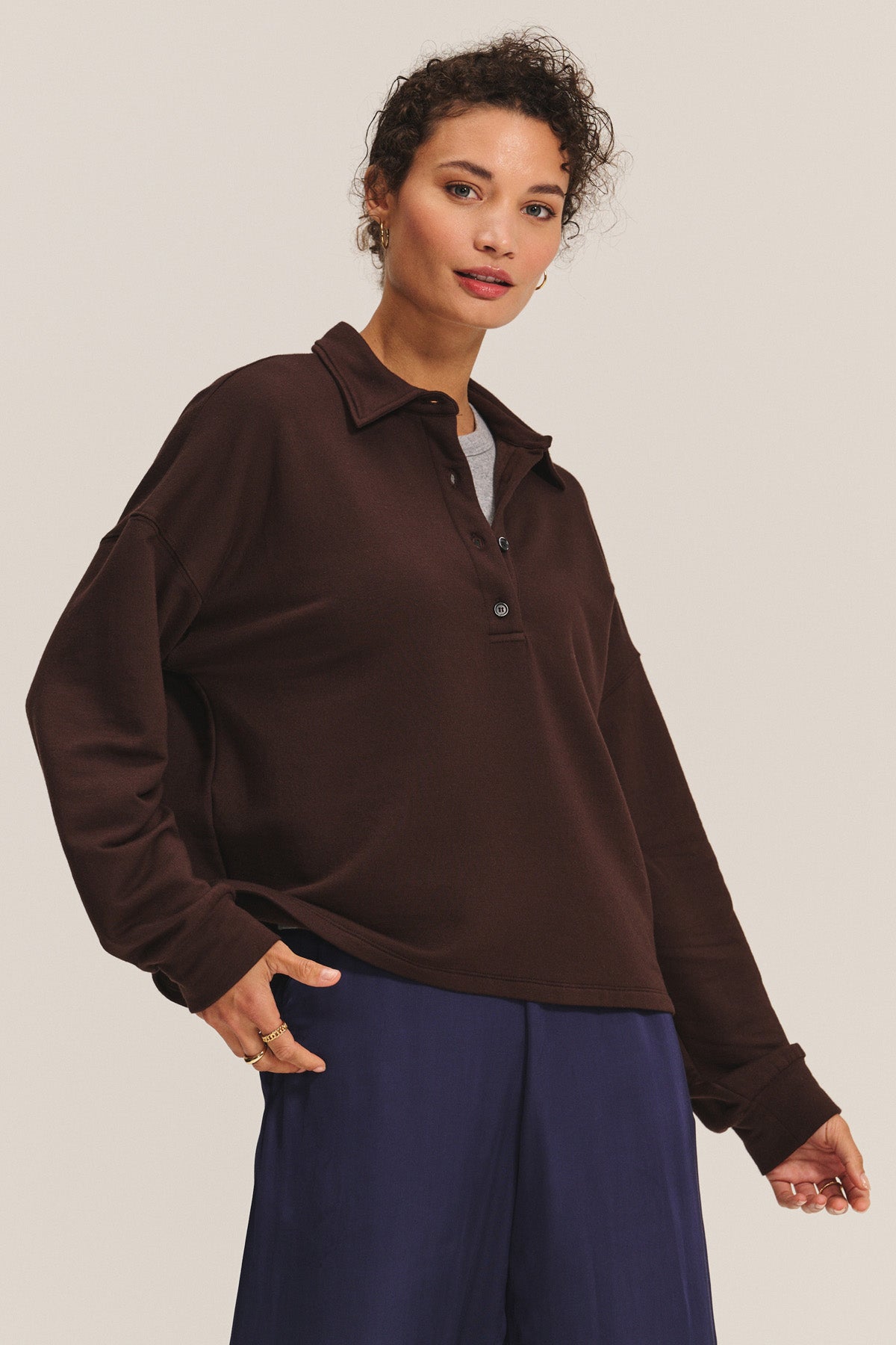 A person wears the MARIAN SWEATSHIRT by Velvet by Graham & Spencer in dark brown over a gray top with navy pants, standing against a light background. The relaxed silhouette creates an effortlessly stylish look.