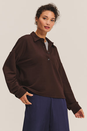A person wears the MARIAN SWEATSHIRT by Velvet by Graham & Spencer in dark brown over a gray top with navy pants, standing against a light background. The relaxed silhouette creates an effortlessly stylish look.