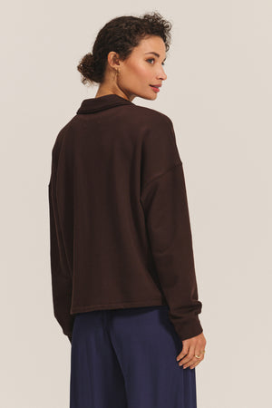 A woman faces away in the Velvet by Graham & Spencer MARIAN Sweatshirt, a dark brown collared top with a relaxed fit, paired with navy pants against a plain light background.
