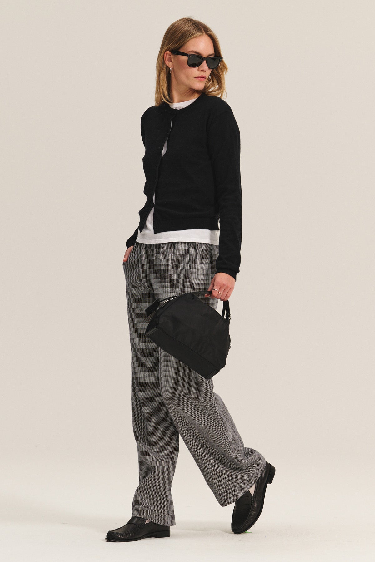 A woman stands against a plain background wearing sunglasses, a black cardigan, white shirt, black shoes, and the BRAYLEN PANT by Velvet by Graham & Spencer in gray cotton gauze with a wide-leg silhouette. She holds a small black bag.-40334106296513