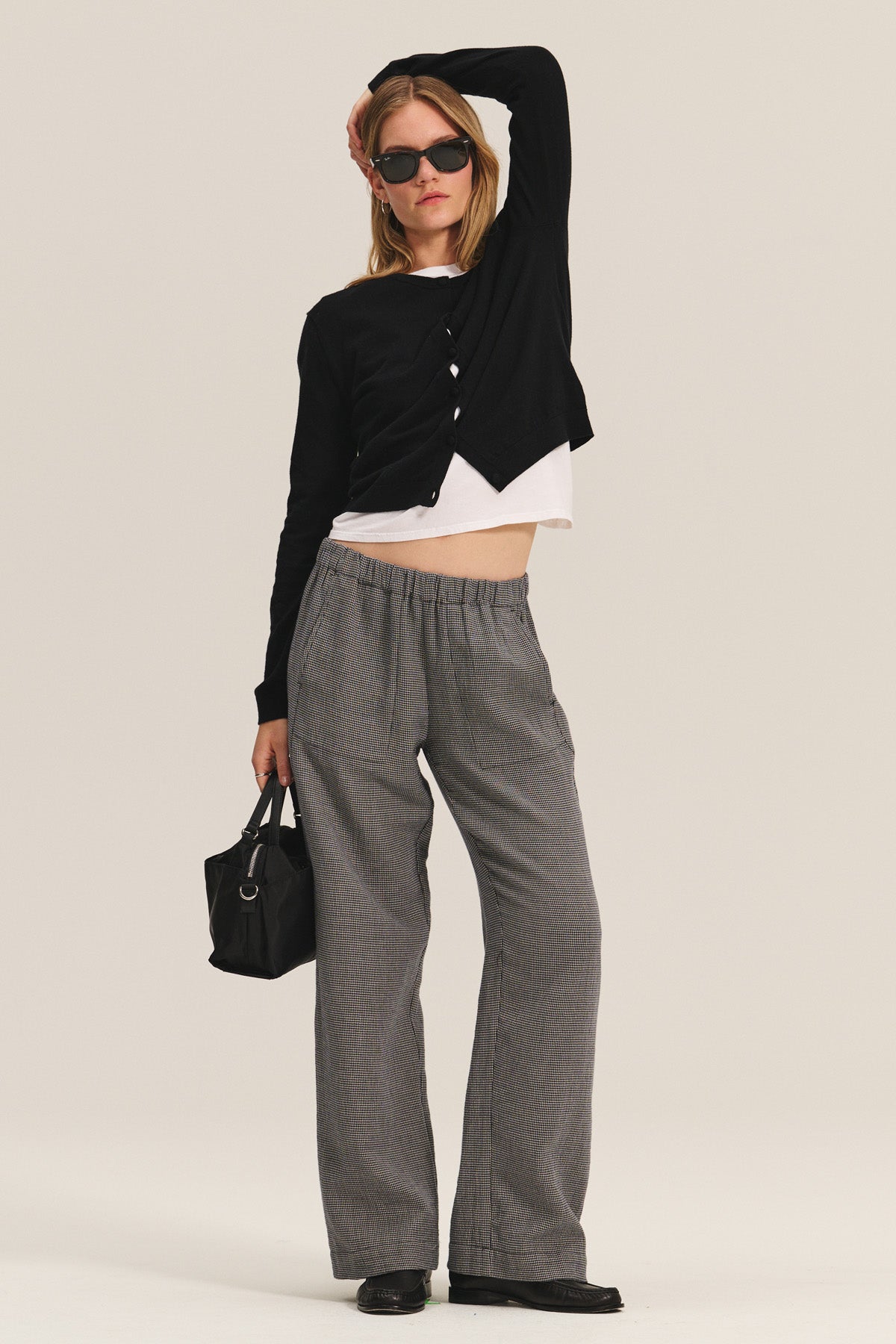 A woman poses against a light background wearing the BRAYLEN PANT from Velvet by Graham & Spencer, paired with a black cardigan, white crop top, black shoes, sunglasses, and holding a black handbag.