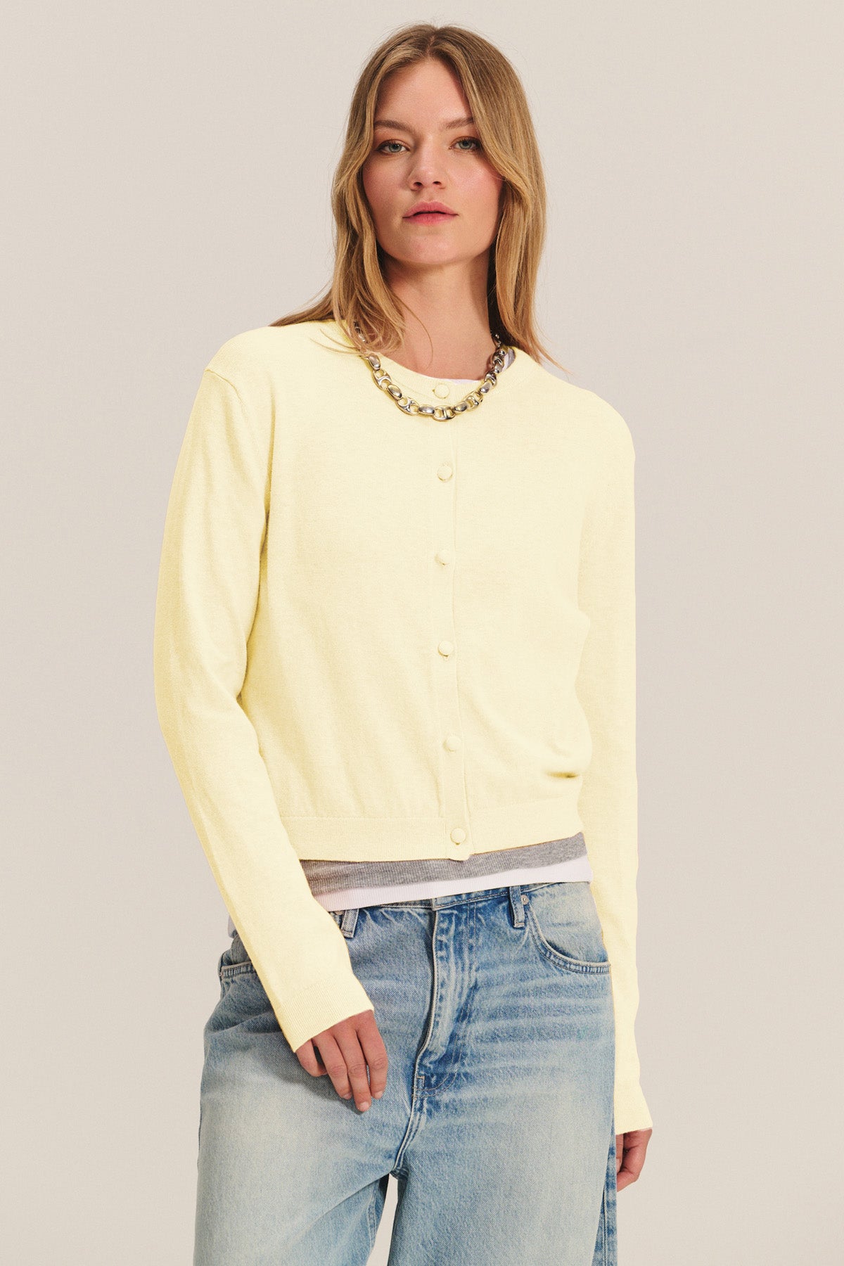 A woman wears the ANGELICA CARDIGAN by Velvet by Graham & Spencer in light yellow over a gray undershirt with blue jeans and a chunky chain necklace, standing against a neutral background.