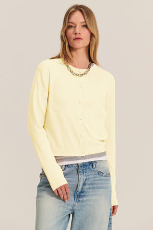 A woman wears the ANGELICA CARDIGAN by Velvet by Graham & Spencer in light yellow over a gray undershirt with blue jeans and a chunky chain necklace, standing against a neutral background.