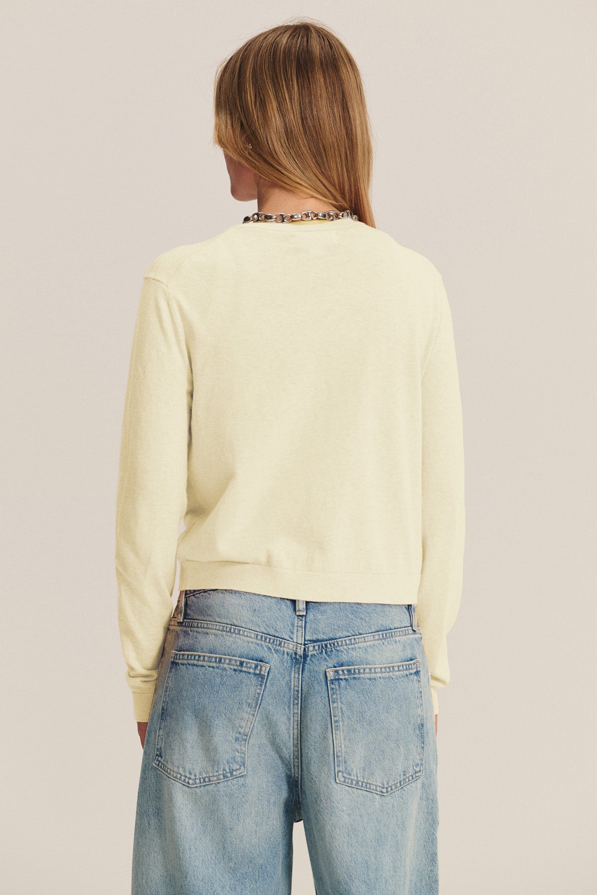A person with long, light brown hair is seen from behind wearing the ANGELICA CARDIGAN by Velvet by Graham & Spencer in light yellow, paired with loose-fitting blue jeans against a plain background.-40732423979201