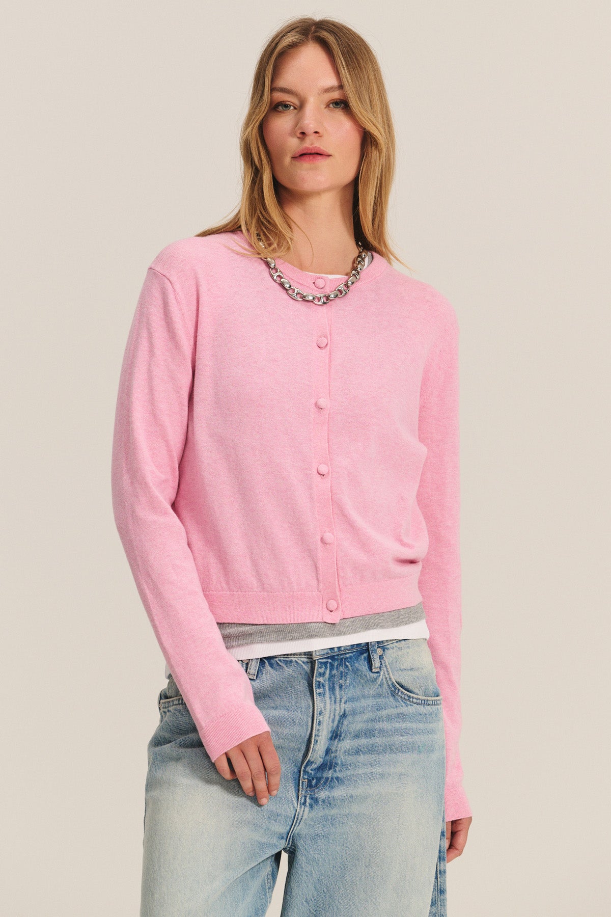 A person wears the ANGELICA CARDIGAN by Velvet by Graham & Spencer in light pink over a gray top and light blue jeans, accessorized with a silver chain necklace.