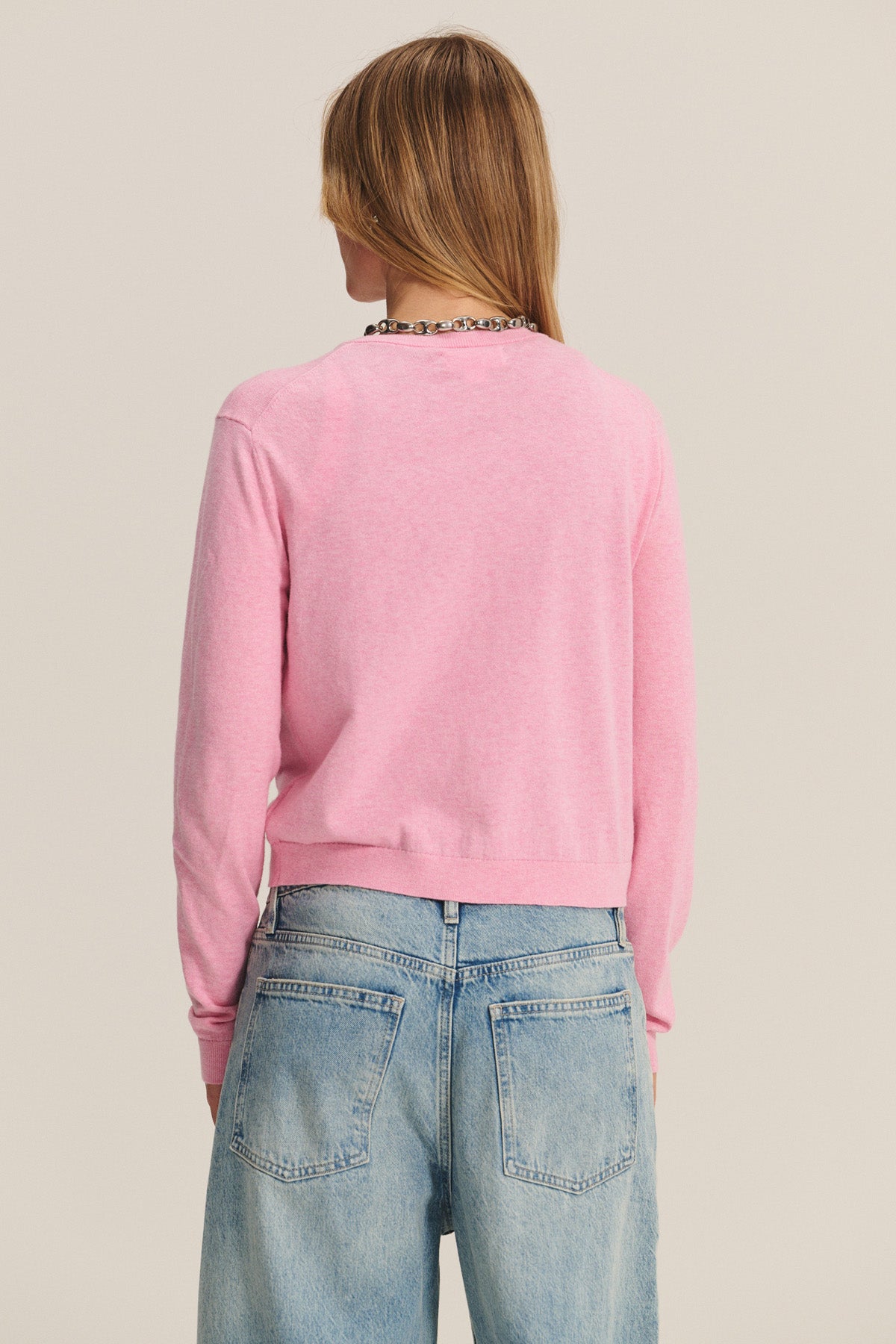 A person with long, light brown hair faces away from the camera, wearing the ANGELICA CARDIGAN by Velvet by Graham & Spencer in pink with light blue jeans.