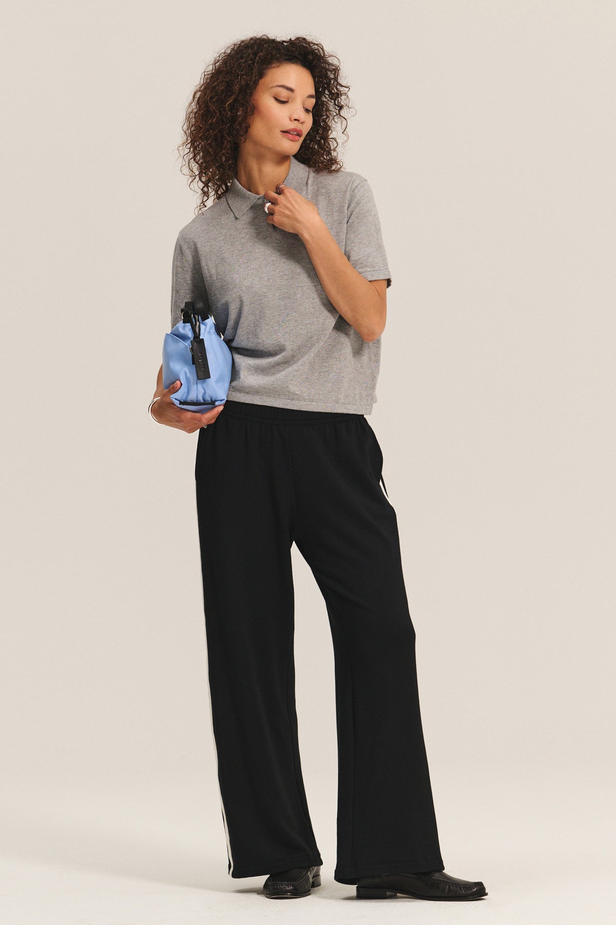 Wearing the EMERSON EMBELLISHED SWEATER POLO by Velvet by Graham & Spencer and black wide-leg pants, a person stands holding a small blue bag with a black strap, showcasing a refined style.-40247631872193