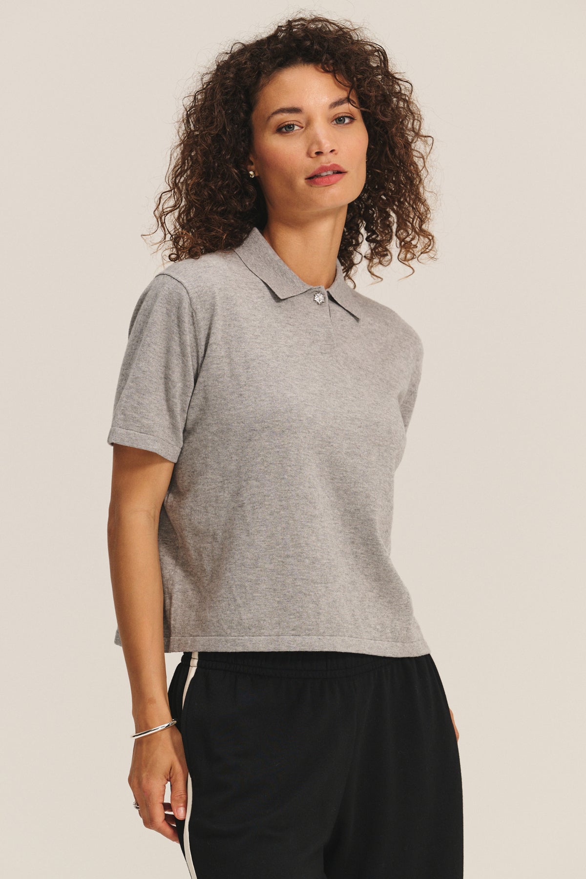 A woman with curly hair models the EMERSON EMBELLISHED SWEATER POLO by Velvet by Graham & Spencer in grey, paired with black pants featuring a white stripe, standing against a plain background.-40247631806657