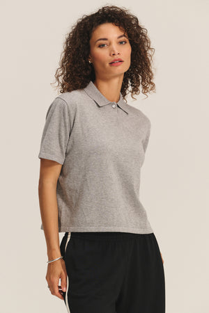 A woman with curly hair models the EMERSON EMBELLISHED SWEATER POLO by Velvet by Graham & Spencer in grey, paired with black pants featuring a white stripe, standing against a plain background.
