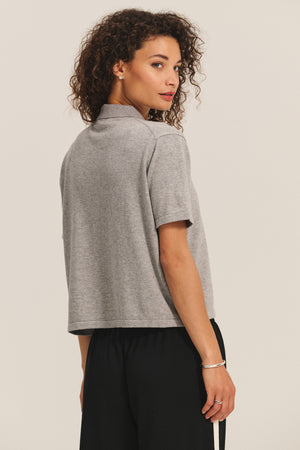 A person with curly hair wears the EMERSON EMBELLISHED SWEATER POLO by Velvet by Graham & Spencer and black pants, standing and looking over their shoulder against a plain background.