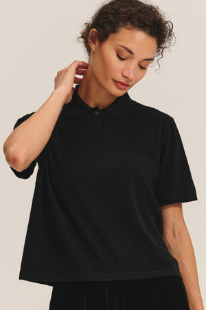 A person wearing the EMERSON SWEATER POLO by Velvet by Graham & Spencer in black stands against a light background, with one hand touching their neck.