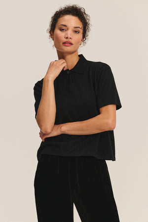 A person wearing the black EMERSON SWEATER POLO by Velvet by Graham & Spencer and black pants stands against a plain light background, touching their chin.