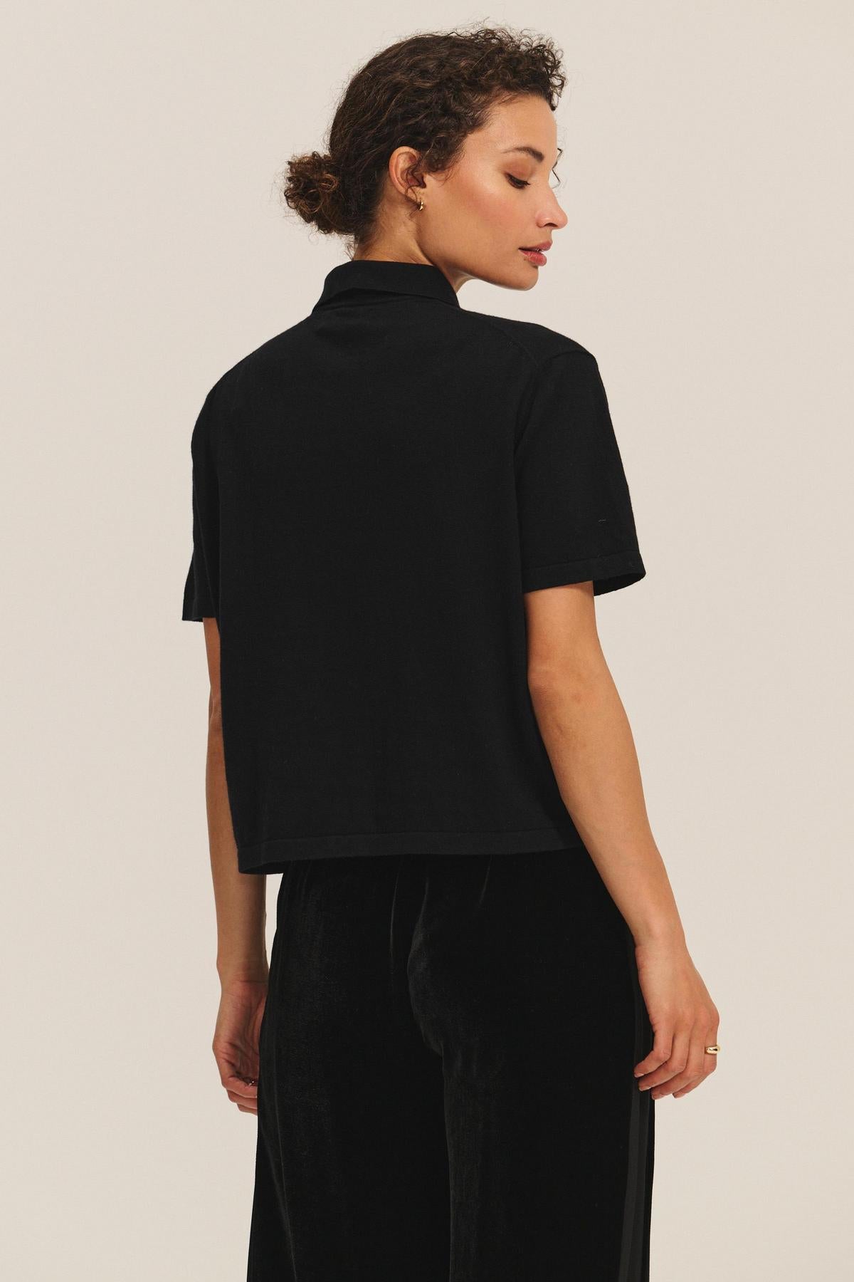 A woman stands with her back to the camera, wearing the black EMERSON SWEATER POLO by Velvet by Graham & Spencer and black pants, set against a plain light background.