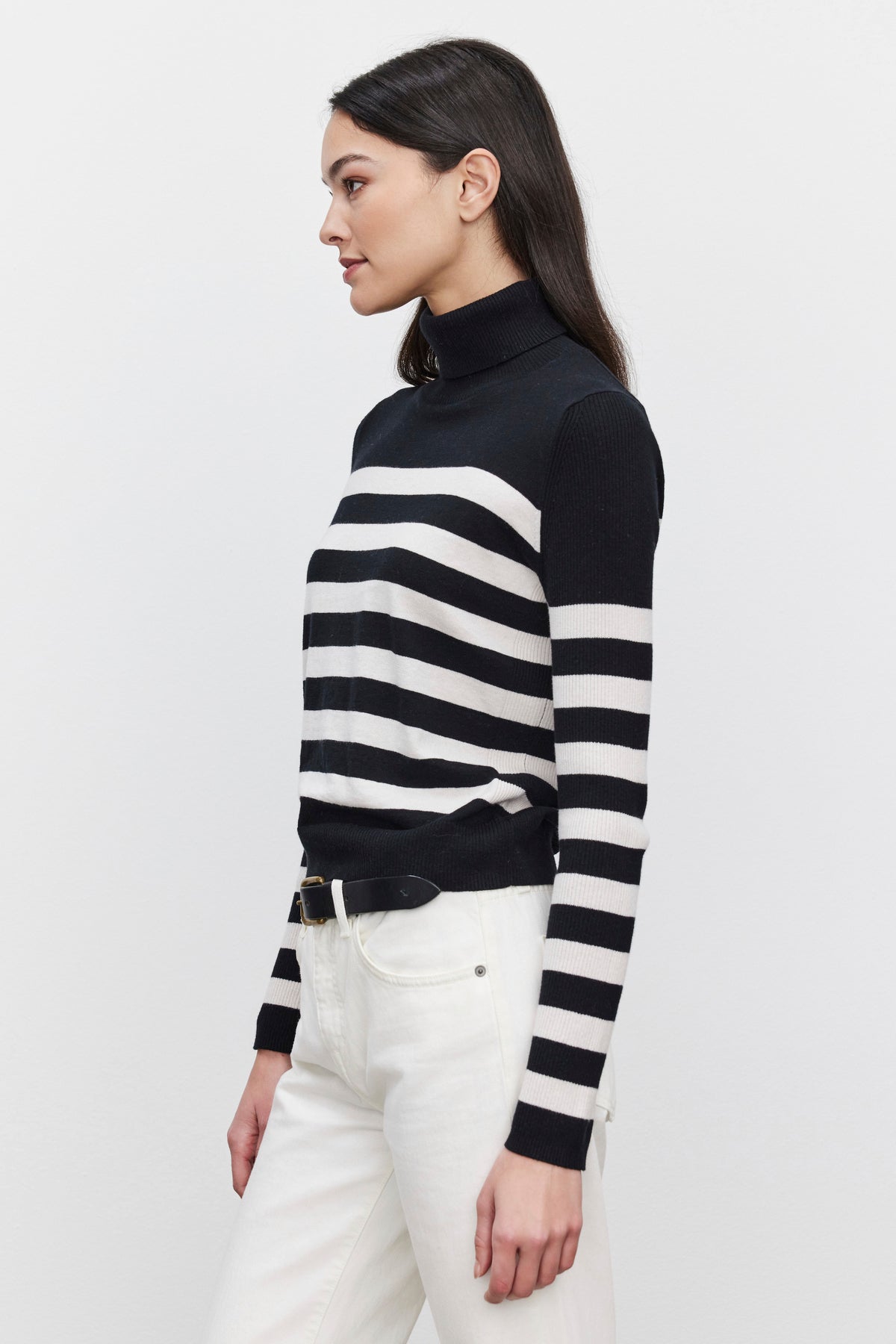   Side profile of a woman wearing the KORIE TURTLENECK SWEATER by Velvet by Graham & Spencer, a black and white striped, fitted turtleneck with ribbed details, paired with white pants, standing against a plain background. 