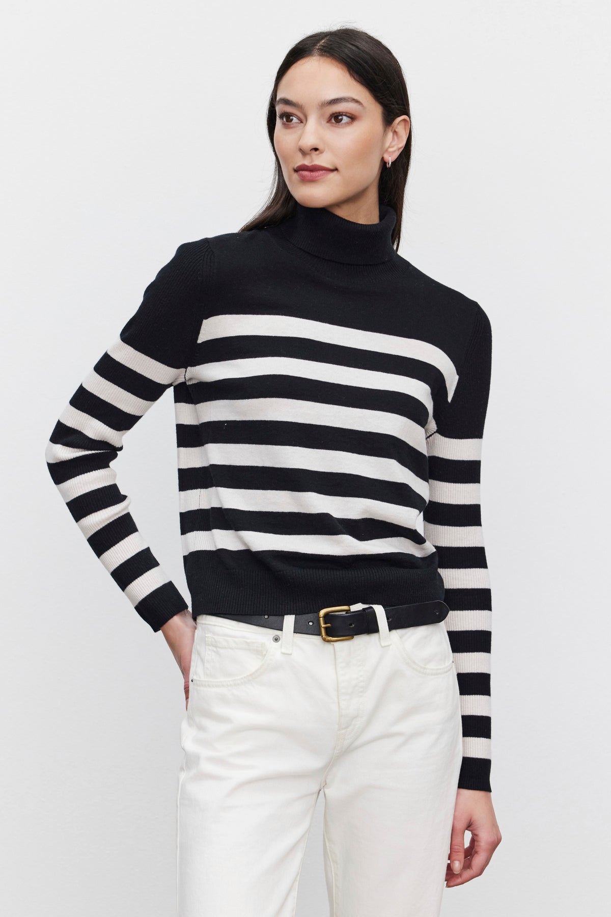   A person is wearing the KORIE TURTLENECK SWEATER by Velvet by Graham & Spencer, which features black and white stripes, a fitted design, and ribbed details. They are also wearing white pants and standing against a plain background. 