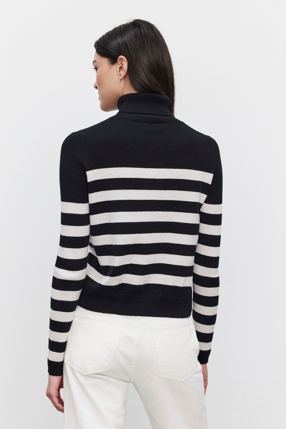   A person is standing with their back to the camera, wearing the KORIE TURTLENECK SWEATER by Velvet by Graham & Spencer and white pants. 