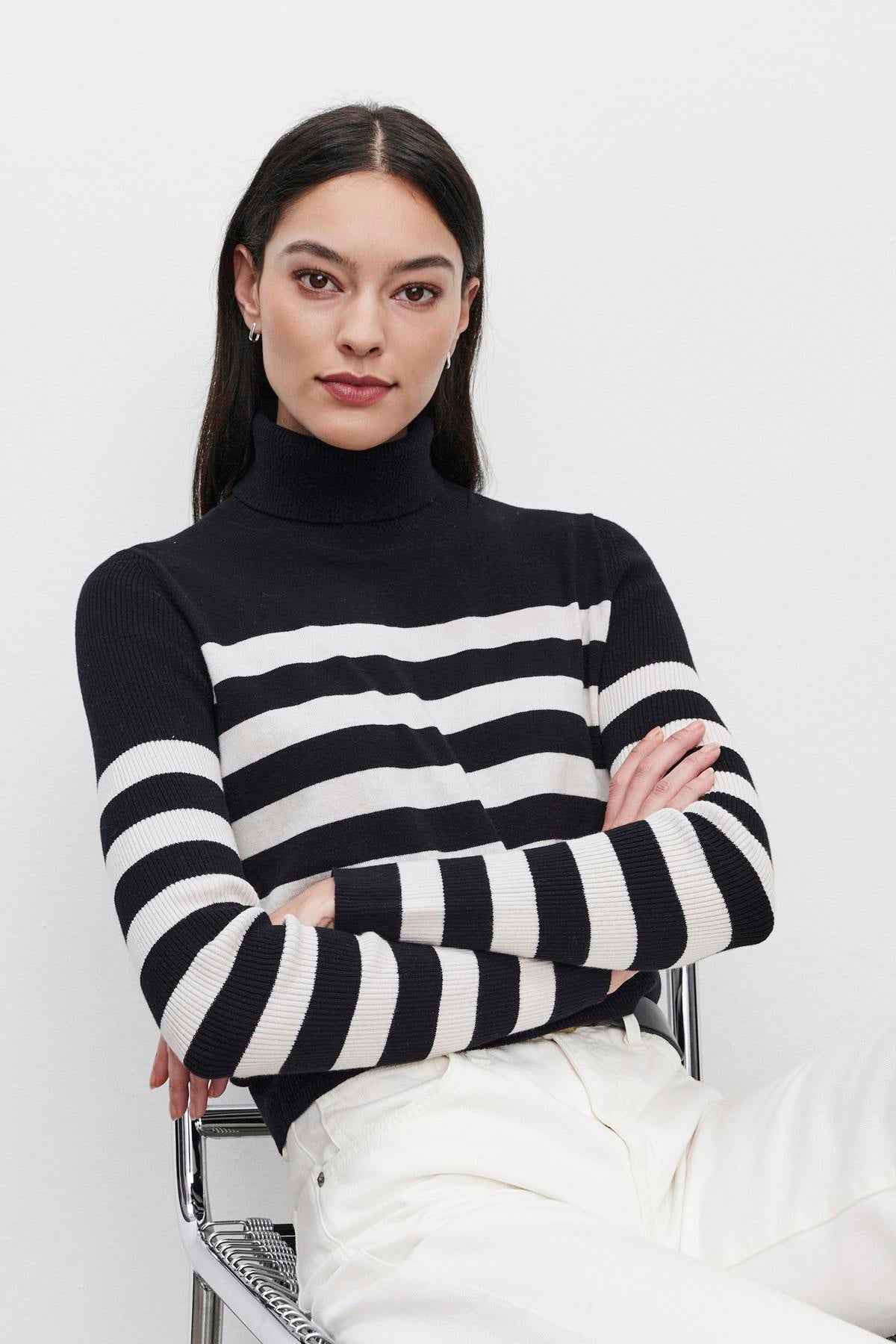   A person with long dark hair sits with arms crossed, wearing the KORIE TURTLENECK SWEATER by Velvet by Graham & Spencer, featuring ribbed details in black and white, paired with white pants, against a plain background. 