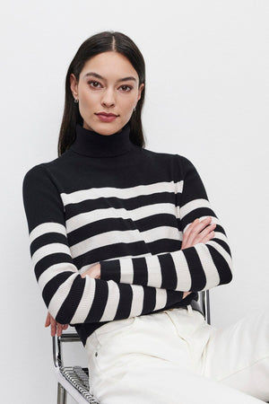 A person with long dark hair sits with arms crossed, wearing the KORIE TURTLENECK SWEATER by Velvet by Graham & Spencer, featuring ribbed details in black and white, paired with white pants, against a plain background.