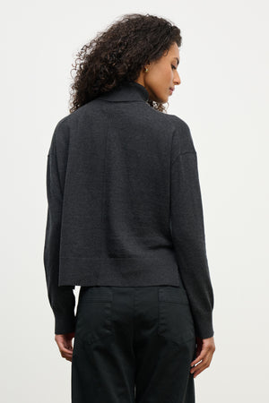 A woman with curly hair wears the LIBRA SWEATER by Velvet by Graham & Spencer, a dark gray cotton cashmere turtleneck in a boxy fit, paired with black pants, shown from the back against a plain light background.